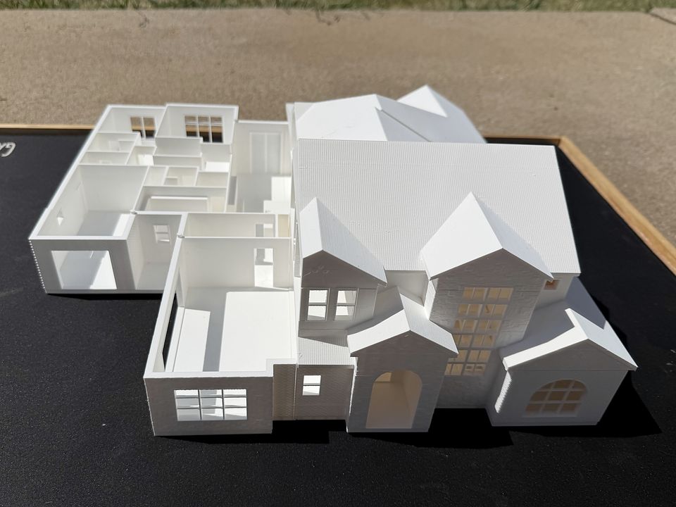 The Adeline by BR Custom Homes: a large white 3D printed two-story custom home model on a black display mat. The roof is lifted off one wing to show the rooms inside, while the front keeps its gables, arched entry and window grids.