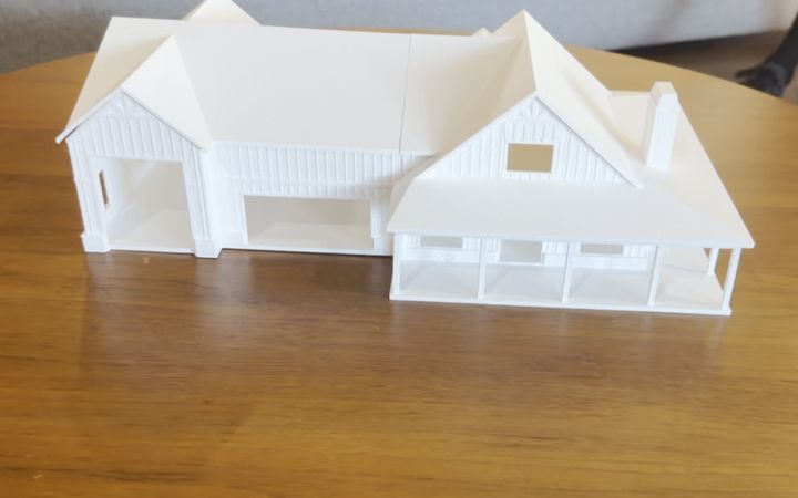 A white 3D printed farmhouse model with a covered porch and chimney on a round wooden table