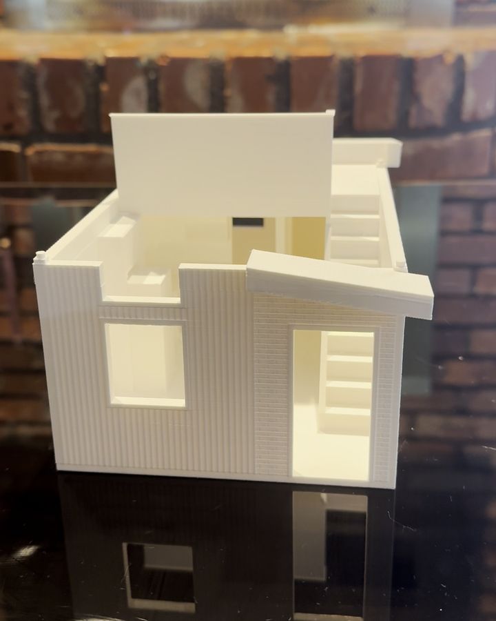 A modern two-story 3D printed model with vertical siding and an awning, opened up on a dark table