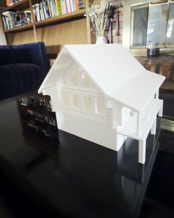A white 3D printed log cabin model with a gabled roof and covered deck