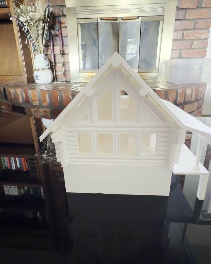 A white 3D printed log cabin model with a gabled roof and side deck, on a glossy black table